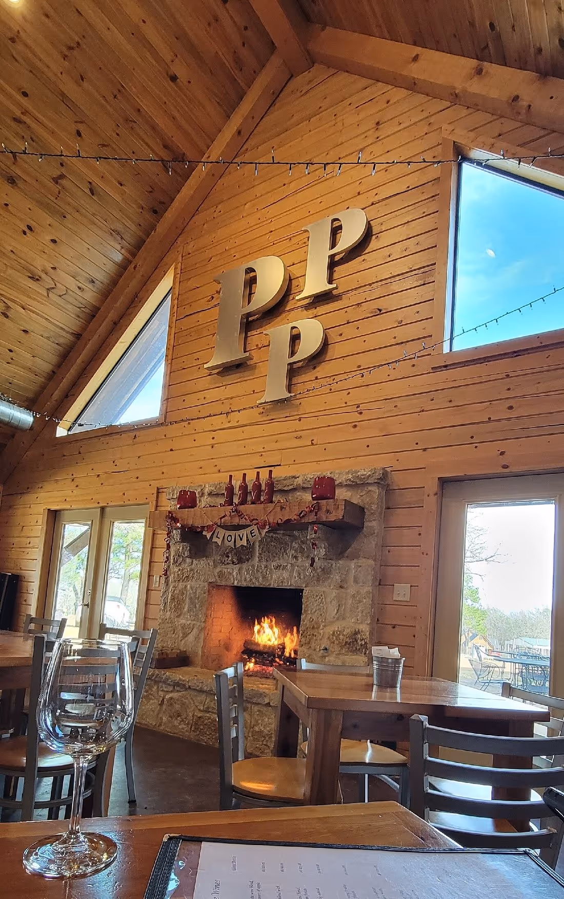 Cozy wooden restaurant interior with stone fireplace, lit fire, tables, chairs, and large Pp letters on the wall.